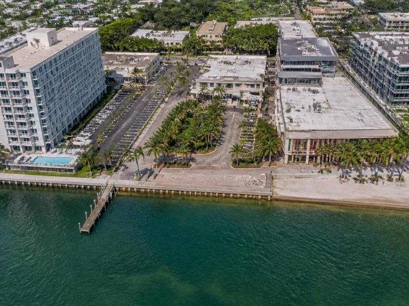Aerial view of a waterfront commercial complex with palm trees, parking lots, and a dock along turquoise water
