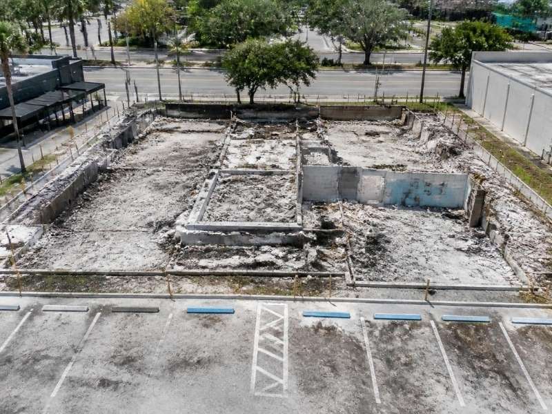 Fire-damaged retail building site with exposed foundations, remaining walls, parking areas, and survey markers in St. Petersburg, Florida.